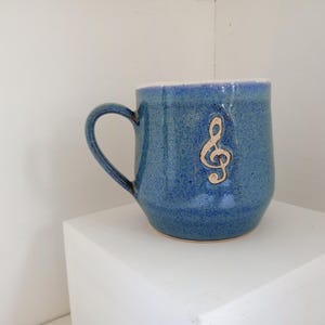 May include: A blue ceramic mug with a gold treble clef design. The mug has a rounded shape with a handle and a white interior. The mug is sitting on a white surface.