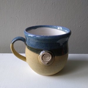 May include: A blue and yellow ceramic mug with a swirl design and a raised circular accent. The mug has a handle and is ideal for enjoying a hot drink.
