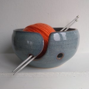 May include: A blue ceramic yarn bowl with a ball of orange yarn and two knitting needles.