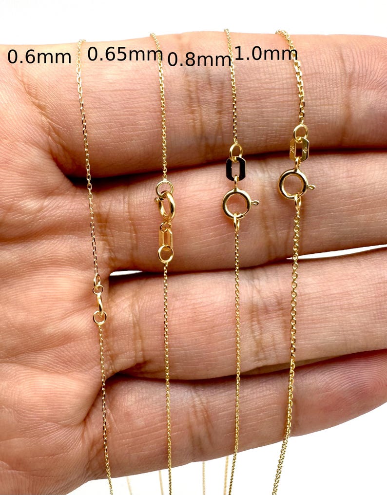 May include: Four gold cable chains with varying thicknesses labeled 0.6mm, 0.65mm, 0.8mm, and 1.0mm. Each chain features a circular spring ring clasp and a small rectangular tag near the closure, showcasing the different link sizes side by side.