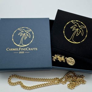 May include: A gold-colored metal chain necklace with a small oval pendant featuring an embossed figure, presented in a dark blue box with a black velvet insert. The box lid displays a gold palm tree logo and the text CARMELFINEFRAFTS 2020.