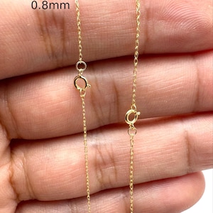 Może przedstawiać: Dwa cienkie złote łańcuszki z zapięciami typu federing, oznaczone tekstem wskazującym na grubości 0,8 mm i 1,0 mm. Delikatne metalowe ogniwa tworzą ciągły wz&oacute;r splotu typu cable, ukazując r&oacute;żnicę w szerokości między tymi dwiema sztukami biżuterii.