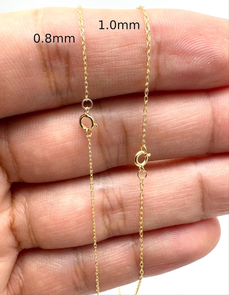 Może przedstawiać: Dwa cienkie złote łańcuszki z zapięciami typu federing, oznaczone tekstem wskazującym na grubości 0,8 mm i 1,0 mm. Delikatne metalowe ogniwa tworzą ciągły wz&oacute;r splotu typu cable, ukazując r&oacute;żnicę w szerokości między tymi dwiema sztukami biżuterii.