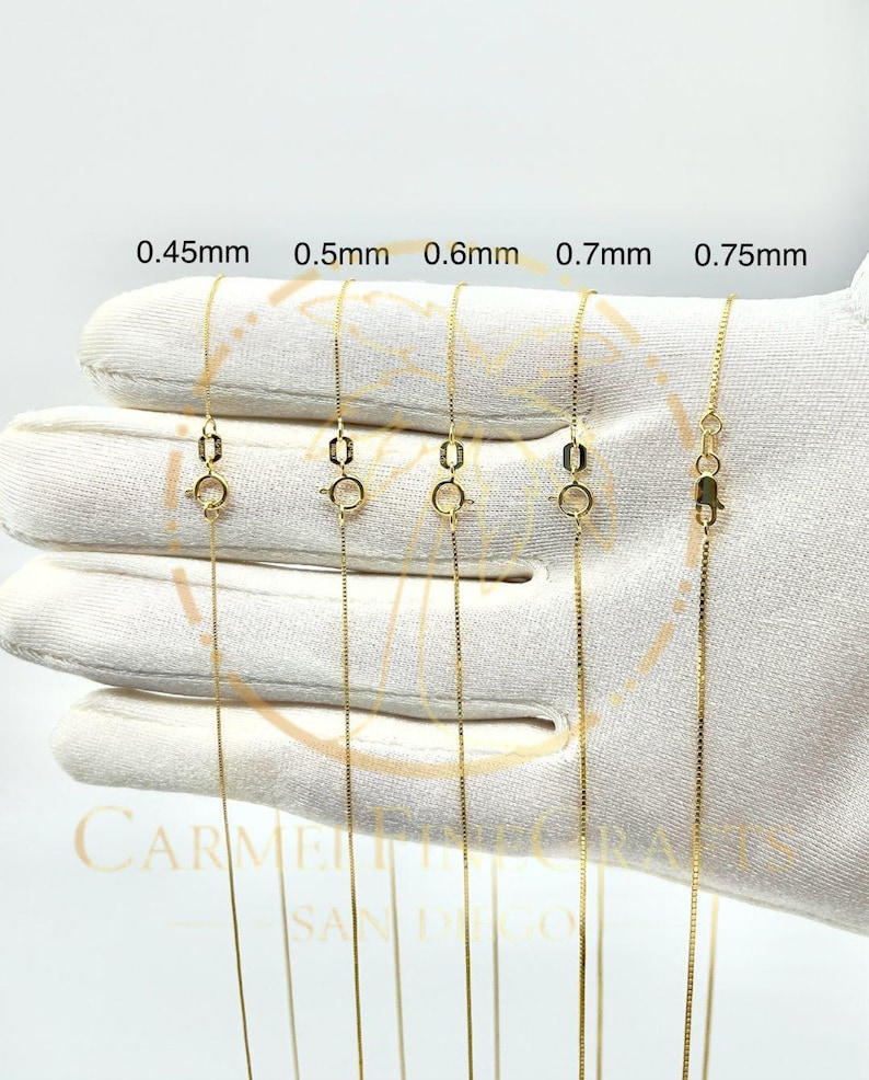 K&ouml;nnte beinhalten: Nahaufnahme von f&uuml;nf goldfarbenen Box-Chain-Halsketten mit unterschiedlichen St&auml;rken, von 0,45 mm bis 0,75 mm. Jede Kette wird mit ge&ouml;ffnetem Verschluss gezeigt, um die Details des Schmucks hervorzuheben. Die Halsketten werden auf einer wei&szlig;en Oberfl&auml;che pr&auml;sentiert.