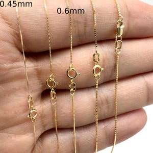 Puede incluir: Cinco cadenas de oro tipo caja con grosores variables etiquetadas como 0,45 mm, 0,5 mm, 0,6 mm, 0,7 mm y 0,75 mm. Cada cadena cuenta con un pequeño cierre de resorte circular y una placa rectangular cerca del cierre, mostrando los anchos.
