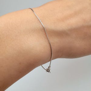 Può includere: Un braccialetto a catena sottile color argento con un delicato design a maglie quadrate. La catena metallica avvolge il polso ed è fissata con una piccola chiusura a moschettone funzionale visibile all'estremità delle maglie.