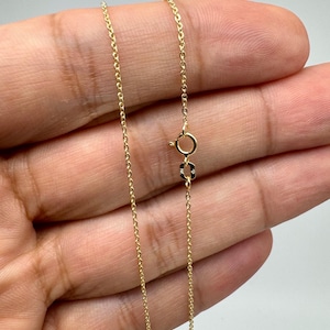 May include: A delicate gold chain necklace featuring a thin cable link design. The necklace is secured with a small, circular spring ring clasp and a rectangular tag attached near the closure, showcasing a polished, reflective metallic finish.