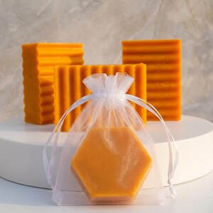 May include: Three rectangular orange bars with a ribbed texture and one hexagonal orange bar inside a sheer white organza drawstring pouch. The items are arranged together, showcasing the vibrant orange color and distinct geometric shapes.