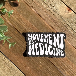May include: A black and white sticker with the words "MOVEMENT IS MEDICINE" in a retro font. The sticker has a scalloped edge and is set against a wooden background with green foliage.