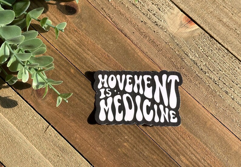 May include: A black and white sticker with the words "MOVEMENT IS MEDICINE" in a retro font. The sticker has a scalloped edge and is set against a wooden background with green foliage.