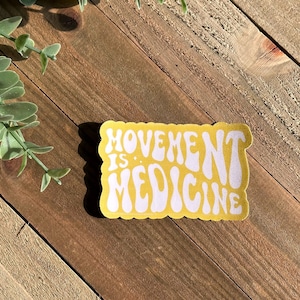 May include: A yellow sticker with the words "MOVEMENT IS MEDICINE" in white, retro lettering. The sticker has a rectangular shape with rounded edges. It is placed on a wooden surface, with some green foliage visible.