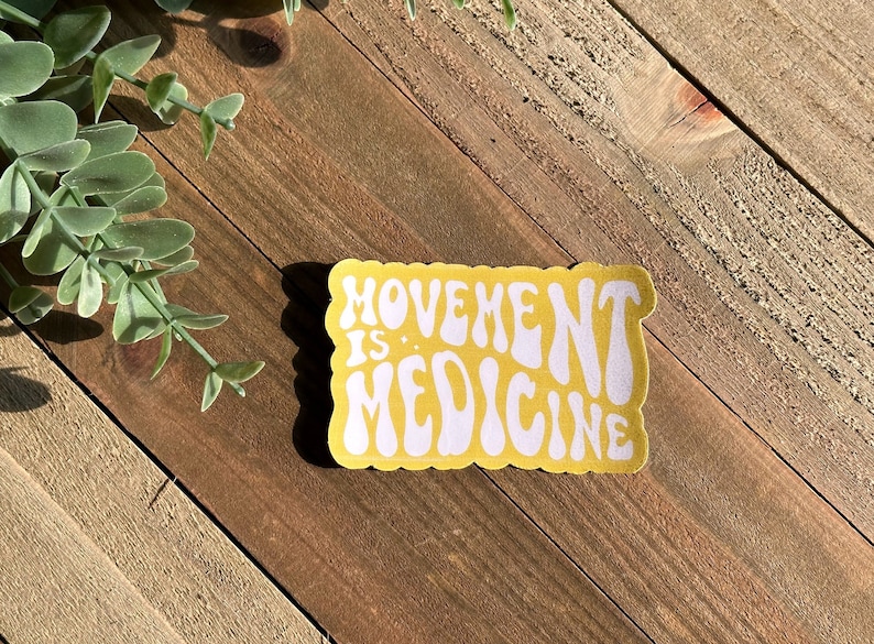 May include: A yellow sticker with the words "MOVEMENT IS MEDICINE" in white, retro lettering. The sticker has a rectangular shape with rounded edges. It is placed on a wooden surface, with some green foliage visible.