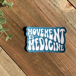 May include: A teal sticker with the words "MOVEMENT IS MEDICINE" in white, retro-style lettering. The sticker has a scalloped edge and is set against a wooden background with a sprig of greenery.