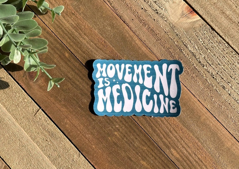 May include: A teal sticker with the words "MOVEMENT IS MEDICINE" in white, retro-style lettering. The sticker has a scalloped edge and is set against a wooden background with a sprig of greenery.