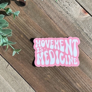 May include: A pink sticker with the words "MOVEMENT IS MEDICINE" in white, retro-style lettering. The sticker is on a wooden surface with a sprig of green leaves in the upper left corner.