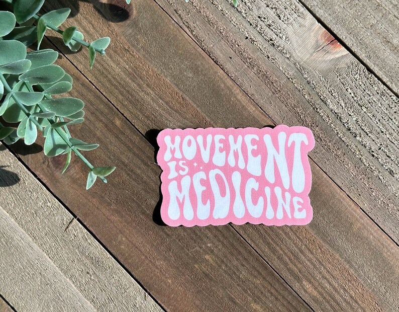 May include: A pink sticker with the words "MOVEMENT IS MEDICINE" in white, retro-style lettering. The sticker is on a wooden surface with a sprig of green leaves in the upper left corner.