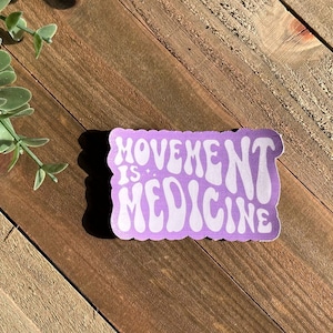 May include: A lavender sticker with the words "MOVEMENT IS: MEDICINE" in white, wavy font. The sticker has a scalloped edge and is set against a wooden background with a sprig of greenery.