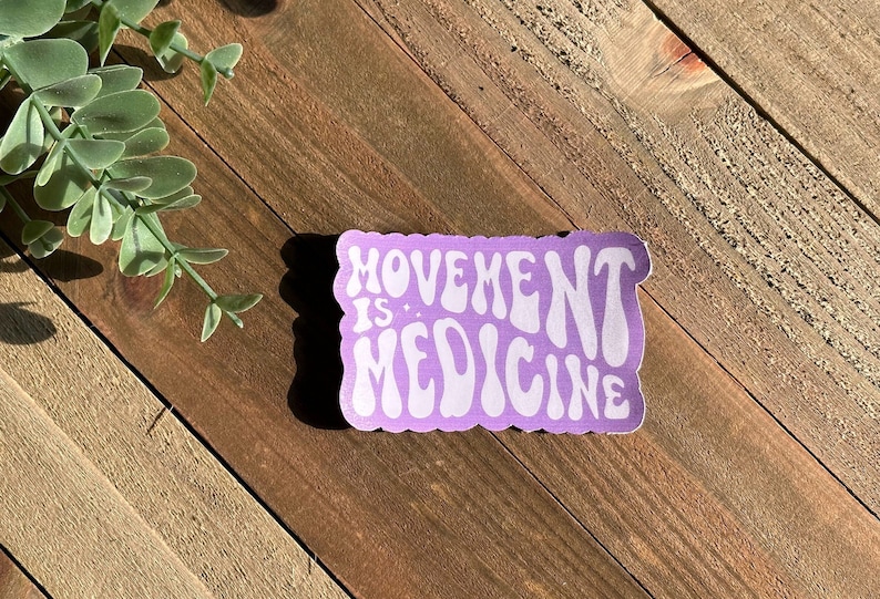 May include: A lavender sticker with the words "MOVEMENT IS: MEDICINE" in white, wavy font. The sticker has a scalloped edge and is set against a wooden background with a sprig of greenery.