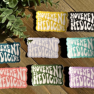 May include: A selection of stickers displaying the text "Movement is Medicine" in a retro style. The stickers are in a range of colours: yellow, black, teal, lavender, pink, green, and purple. They are arranged on a wooden surface.