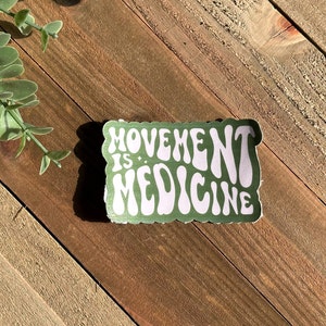 May include: A rectangular sticker with the words "MOVEMENT IS MEDICINE" in white, retro-style font, on a green background. The sticker is on a wooden surface, with a sprig of greenery in the upper left corner.