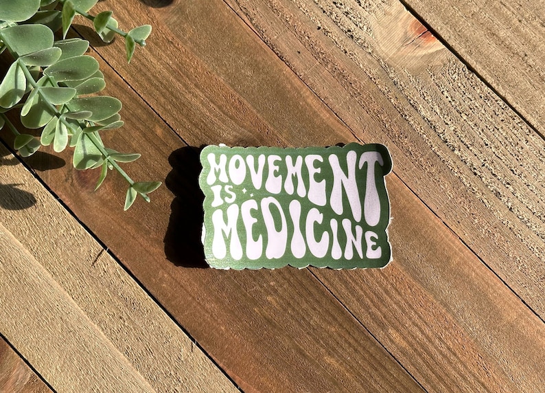 May include: A rectangular sticker with the words "MOVEMENT IS MEDICINE" in white, retro-style font, on a green background. The sticker is on a wooden surface, with a sprig of greenery in the upper left corner.