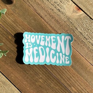 May include: A rectangular sticker with the words "MOVEMENT IS MEDICINE" in white, retro-style font. The sticker has a light blue background and a scalloped edge. The sticker is on a wooden surface.