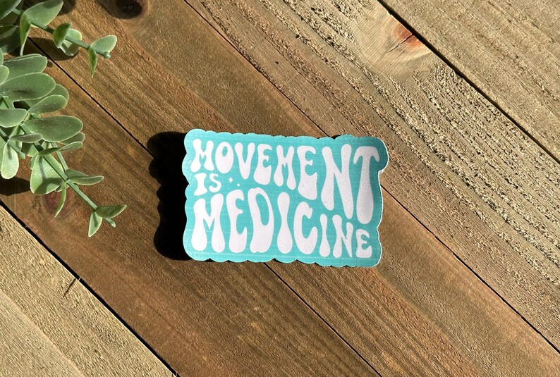 May include: A rectangular sticker with the words "MOVEMENT IS MEDICINE" in white, retro-style font. The sticker has a light blue background and a scalloped edge. The sticker is on a wooden surface.