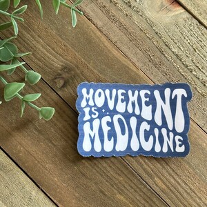 May include: A blue sticker with white text that reads "MOVEMENT IS MEDICINE." The sticker has a wavy, retro font and a white border. The sticker is on a wooden surface with green foliage in the background.