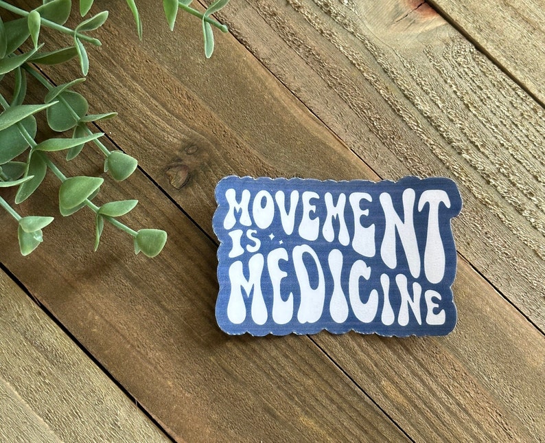 May include: A blue sticker with white text that reads "MOVEMENT IS MEDICINE." The sticker has a wavy, retro font and a white border. The sticker is on a wooden surface with green foliage in the background.