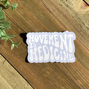 May include: A light blue sticker with the words "MOVEMENT IS MEDICINE" in white, retro-style lettering. The sticker is rectangular with rounded edges and is placed on a wooden surface.