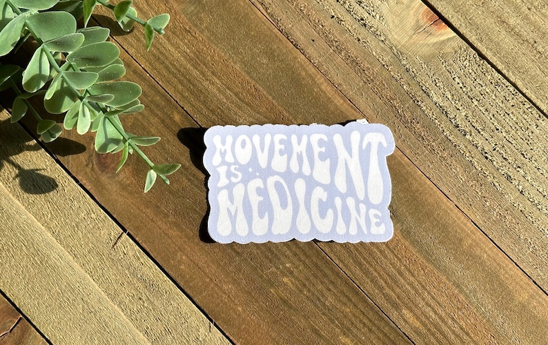 May include: A light blue sticker with the words "MOVEMENT IS MEDICINE" in white, retro-style lettering. The sticker is rectangular with rounded edges and is placed on a wooden surface.