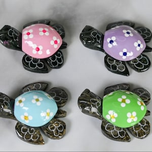 May include: Four decorative turtle figurines with painted shells. The shells are painted in pink, purple, light blue, and green, each adorned with white and yellow flower designs. The turtles' bodies are dark with etched details.