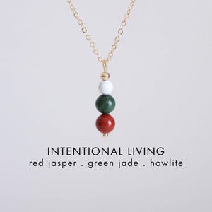 Pu&ograve; includere: Una collana con catena color oro con un ciondolo con tre perline di pietre preziose: diaspro rosso, giada verde e howlite. La collana ha le parole "INTENTIONAL LIVING" e i nomi delle pietre in testo bianco.
