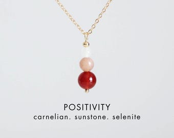 Puede incluir: Un collar de cadena dorada con un colgante vertical de tres cuentas apiladas: una cuenta de selenita blanca, una cuenta de piedra de sol color melocotón y una cuenta de cornalina roja oscura. Texto: POSITIVITY carnelian. sunstone. selenite. CRYSTALLIUM.
