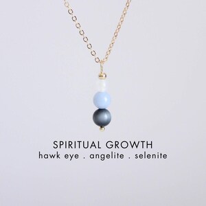 Peut inclure: Un collier à chaîne dorée avec un pendentif vertical composé de trois perles empilées : un œil de faucon gris foncé, une angélite bleu clair et une sélénite blanche. Le texte indique : SPIRITUAL GROWTH, hawk eye . angelite . selenite, CRYSTALLIUM.