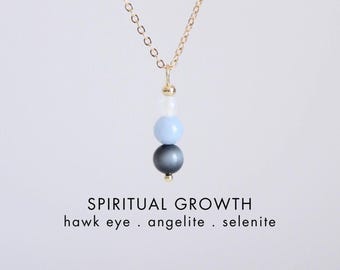 May include: A gold chain necklace featuring a vertical pendant with three stacked beads: a dark grey hawk eye, a light blue angelite, and a white selenite. Text below reads: SPIRITUAL GROWTH, hawk eye . angelite . selenite, and CRYSTALLIUM.