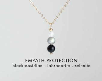 May include: A gold-toned chain necklace featuring a vertical pendant of three round beads: a white selenite bead, a grey labradorite bead, and a black obsidian bead. Text below reads: EMPATH PROTECTION black obsidian . labradorite . selenite CRYSTALLIUM.
