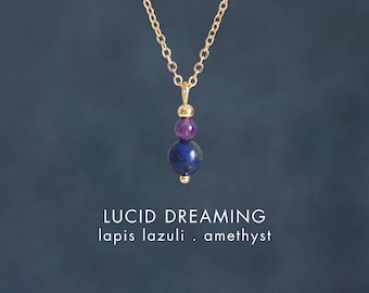 May include: A gold chain necklace featuring a pendant with a small gold bead, a round purple amethyst bead, and a larger round blue lapis lazuli bead. The text reads LUCID DREAMING lapis lazuli . amethyst and the brand name CRYSTALLIUM.