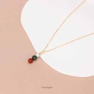 Pu&ograve; includere: Una delicata collana d'oro con un pendente con tre perline di pietre preziose colorate: rosso, verde e bianco. La collana &egrave; esposta su una superficie bianca, su uno sfondo rosa tenue. La parola "CRYSTALLIUM" &egrave; stampata in basso.