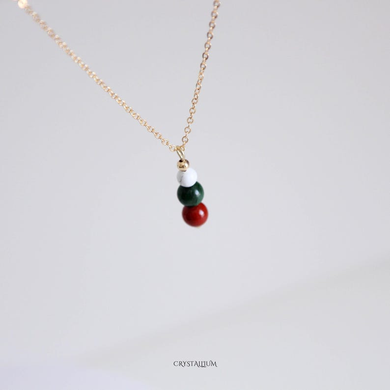 Pu&ograve; includere: Una delicata collana d'oro con un ciondolo composto da quattro perline colorate: rosse, verdi, bianche e dorate. Le perline sono infilate insieme, creando un design semplice ma elegante. La collana &egrave; esposta su uno sfondo morbido e neutro.