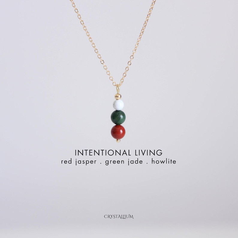 Pu&ograve; includere: Una collana con catena color oro con un ciondolo con tre perline di pietre preziose: diaspro rosso, giada verde e howlite. La collana ha le parole "INTENTIONAL LIVING" e i nomi delle pietre in testo bianco.