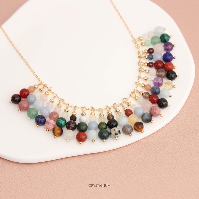 Pu&ograve; includere: Una collana dorata con una cascata di perline di pietre preziose colorate. Le perline sono di varie dimensioni e colori, tra cui rosso, blu, verde e viola. La collana &egrave; esposta su una superficie bianca con la scritta "CRYSTALLIUM" in basso a destra.