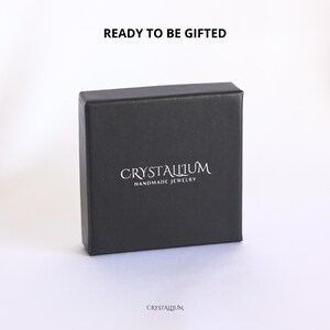 Pu&ograve; includere: Una scatola regalo quadrata nera con la scritta "READY TO BE GIFTED" in alto. La scatola ha il marchio "CRYSTALLIUM" e le parole "HANDMADE JEWELRY" stampate in bianco. Il marchio &egrave; stampato anche in basso.