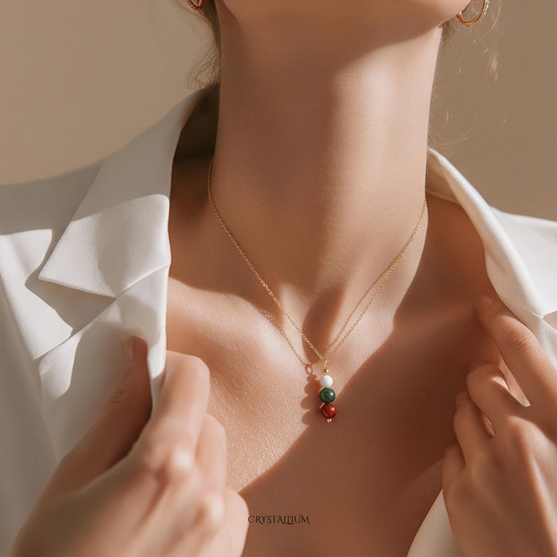 Pu&ograve; includere: Una collana color oro con un pendente composto da tre perle di pietre preziose: bianca, verde e rossa. La collana &egrave; indossata su una camicia bianca. La parola "CRYSTALIUM" &egrave; stampata in basso nell'immagine.