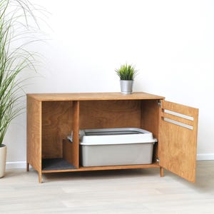 May include: A rectangular wooden cabinet with a light brown wood grain finish, featuring a hinged door with two horizontal slats. Inside, a gray and white plastic litter box sits on the right, while a dark gray mat lines the left side.