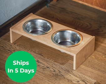 May include: A wooden pet feeder with two stainless steel bowls. The feeder is made of light-colored wood and has a rectangular base. The bowls are round and sit side-by-side. The text "Ships In 5 Days" is in a green circle.