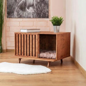 May include: A rectangular wooden pet crate with a sliding slatted door and four short tapered legs. The interior contains a soft, light-coloured cushion. The crate features a warm brown wood grain finish and a clean, boxy structure for small animals.