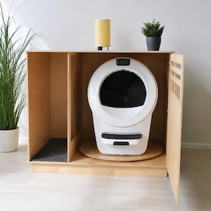 May include: A light-colored wooden cabinet enclosure featuring an open side compartment with a dark gray textured mat and a main section housing a white and black automatic cat litter box. The cabinet has a hinged door and a smooth finish.