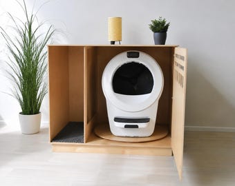 May include: A light-colored wooden cabinet enclosure featuring an open side compartment with a dark gray textured mat and a main section housing a white and black automatic cat litter box. The cabinet has a hinged door and a smooth finish.