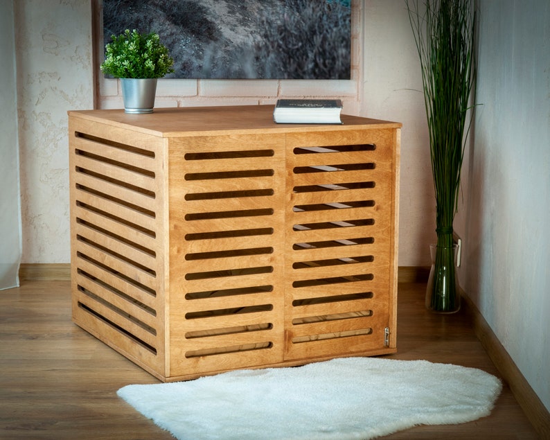 Op de afbeelding: Een rechthoekige houten bench voor huisdieren met een lichtbruine afwerking, voorzien van horizontale latten aan de voor- en zijkanten. De constructie heeft een massief bovenoppervlak en een kleine metalen sluiting in de rechterbenedenhoek van het voorpaneel.